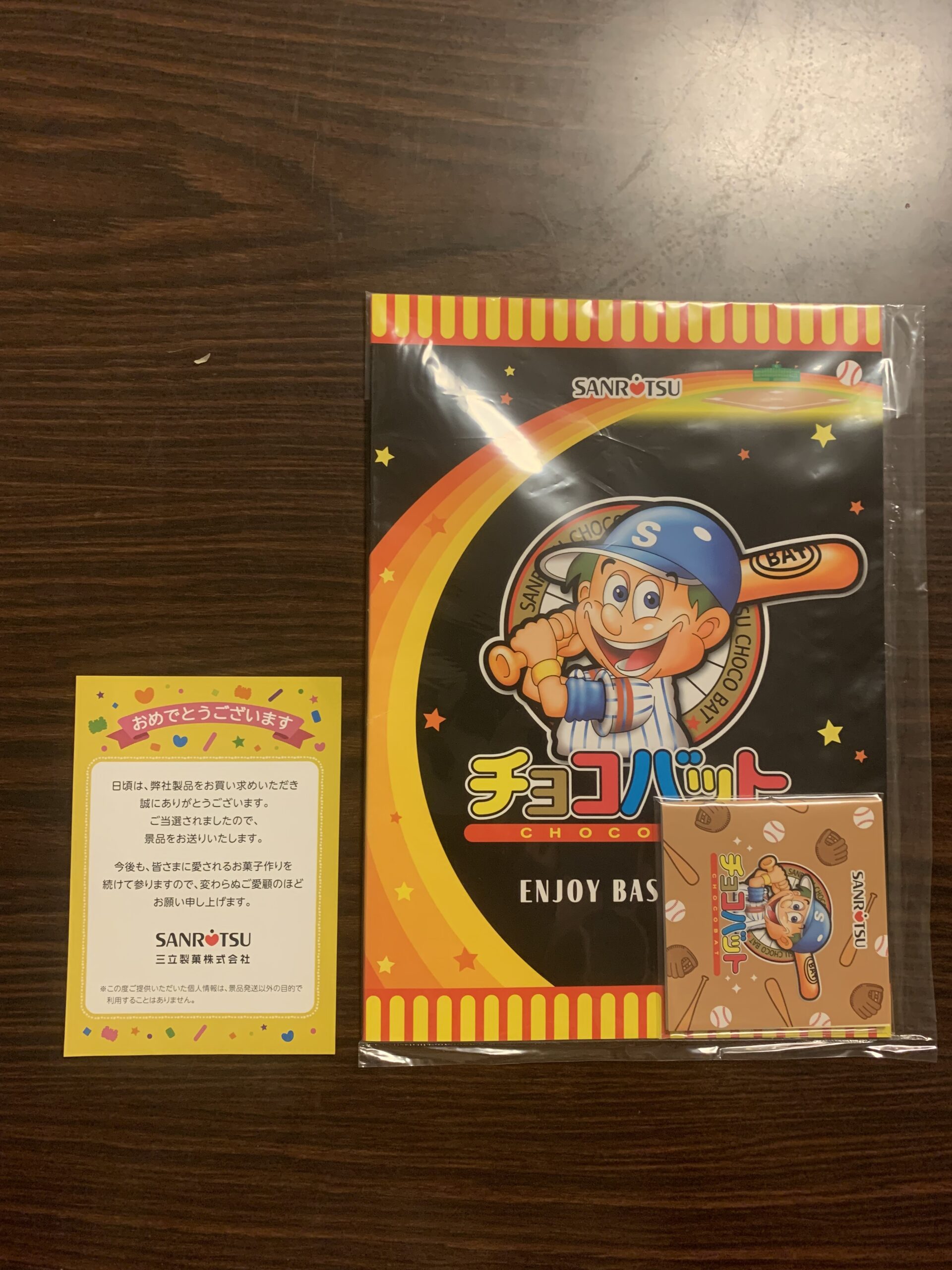 Prize merchandise from Sanritsu Confectionery's "Choco Bat" campaign displayed on a wooden table. The set includes a plastic clear folder and a small notepad featuring the iconic baseball boy mascot, alongside a yellow letter with Japanese text saying "Congratulations". / 木目のテーブルの上に置かれた三立製菓「チョコバット」のキャンペーン当選品。野球少年のマスコットキャラクターが描かれた黒とオレンジのクリアファイル、正方形のメモ帳、そして「おめでとうございます」と書かれた黄色い当選通知カードの3点セット。